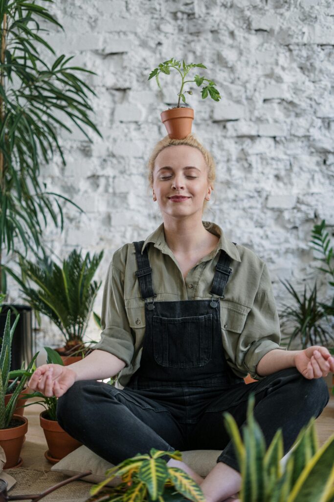 Woman meditating indoors surrounded by potted plants, balancing a pot on her head.